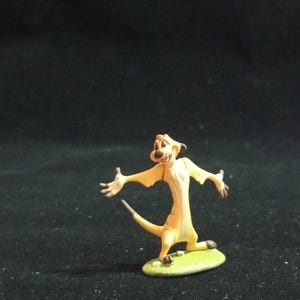 May include: A small, yellow figurine of Timon from The Lion King. The meerkat figure is in a dynamic pose with arms outstretched, standing on a small green base. The figurine has a smiling expression and a long tail. The background is dark.