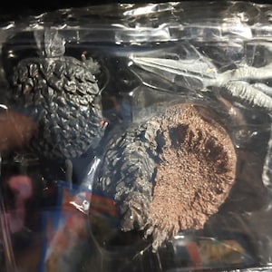 May include: A packaged action figure featuring a detailed, textured, dark gray creature with a brown base. The figure is encased in a clear plastic container, with a black background. The creature has multiple appendages.