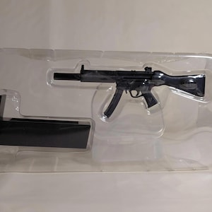 Puede incluir: Una ametralladora de juguete negra y un soporte negro se presentan dentro de un paquete de plástico transparente. El arma presenta detalles intrincados, incluyendo un cargador, un mango y una culata. El soporte es rectangular e inclinado.