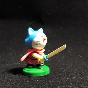 Op de afbeelding: Een klein beeldje van een blauw-wit katachtig personage met een rode cape en een houten zwaard. Het figuur staat op een groene basis. Het personage heeft een blauwe kop en oren, een wit gezicht en een rode outfit.