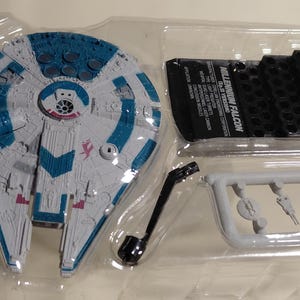 May include: A Millennium Falcon model kit in a clear plastic package. The spaceship is primarily gray with blue and red accents. The kit includes additional black and gray parts, and the text "MILLENNIUM FALCON" is visible on a black label.