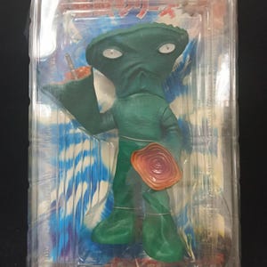 May include: A green alien action figure in a clear plastic package. The figure has large eyes, a wide head, and is holding a small drink. It also has a circular orange object attached to its body. The packaging has Japanese text and the brand name "unifive".