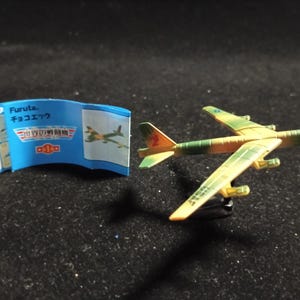 Op de afbeelding: Een miniatuur speelgoedvliegtuig met een camouflagepatroon in groen en geel, met oranje accenten op de staart. Een blauw papier met Japanse tekst ligt naast het vliegtuig.