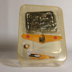 以下が含まれることがあります： 透明なプラスチックパッケージに入った模型飛行機のキット。キットには、赤い円が付いた黄色の翼、胴体、オリーブグリーンのプラスチック部品のスプルーが含まれています。このモデルはヴィンテージ航空機です。