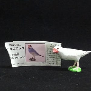 以下が含まれることがあります： 赤い嘴と足を持つ白い鳥の置物。緑色の台座の上に立っています。日本語のテキストと鳥のイラストが描かれた小さなカードが置物の隣にあります。「Furuta」のブランド名がカードに表示されています。