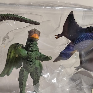 May include: A set of dinosaur toys, including a green and brown dinosaur figure with wings, a dark blue pterodactyl, and a segmented tail. The toys are packaged in a clear plastic container.