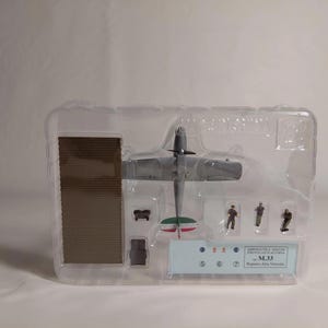 Op de afbeelding: Grijs modelvliegtuig met groen-witte accenten op de staart, gepresenteerd in een transparante plastic verpakking. De set bevat miniatuurfiguren en een bruine rechthoekige structuur. De tekst "AEROMODELLI & MACCHI" is zichtbaar.