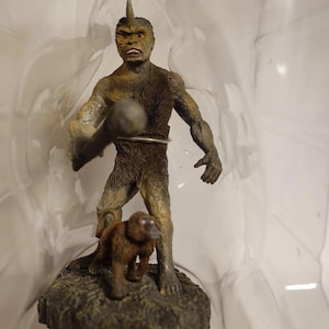 Peut inclure: Figurine détaillée d'une créature avec une corne, tenant une arme et un petit animal à ses pieds. La figurine est principalement marron et beige, avec un aspect texturé. La base est une surface rocheuse sombre.