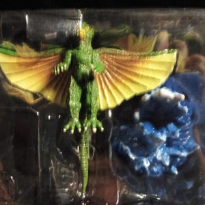 May include: A green, winged creature figurine with yellow and brown wings, packaged in a clear plastic container. A blue, textured element is also included in the packaging.