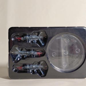May include: A black plastic tray containing three miniature toy fighter jets and a clear plastic display stand. The jets are primarily dark blue with red and silver accents. The tray is on a light background.