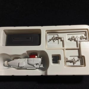 May include: A model submarine kit presented in a white plastic tray. The kit contains a grey submarine model with a red top, a black rectangular component, and various small metal parts. The tray is set against a black backdrop.