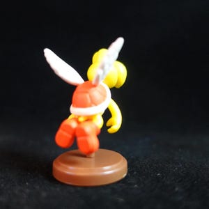 May include: A small figurine of a Koopa Paratroopa, a character from the Super Mario video game series. The figure is yellow with a red shell, orange shoes, and white wings. It is mounted on a brown base.