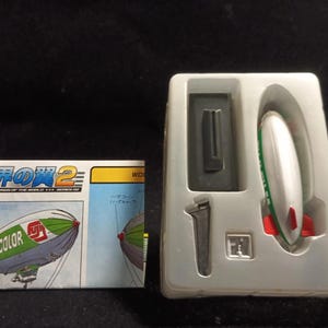 May include: A model kit featuring a white, green, and red blimp with the text "FUJICOLOR" on the side. The kit includes a blimp model, a small gray rectangular piece, and a silver-colored piece, all in a white plastic tray. The box has Japanese text.