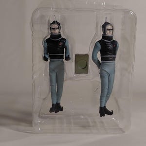 May include: Two figurines of a futuristic character in a clear plastic package. The figures wear black vests, light blue pants, and black boots. They have helmets with visors. A small, green, rectangular piece is between the figures.