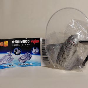 May include: A silver toy spaceship is partially visible, packaged in clear plastic. A black stand and a clear plastic arc are also present. A card with Japanese text and illustrations of toy vehicles is to the left.