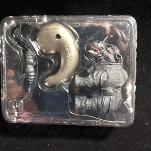 May include: A clear plastic package containing two small figurines. One is a gray monster with red teeth, and the other is a gold-colored, abstract shape. The package is on a dark surface.