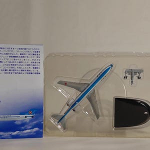 May include: A model airplane, silver with blue accents, displayed in a clear plastic case. The model includes a stand and additional parts. A card with Japanese text and an illustration of the plane is visible.