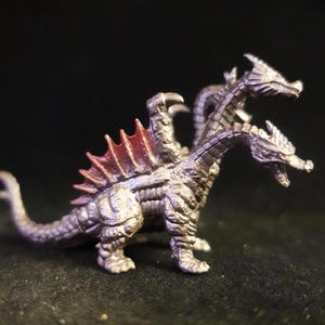 Può includere: Statuetta di drago argentato a tre teste con pinne dorsali rosse. La scultura dettagliata ha una lunga coda ed è in posa sulle quattro zampe. Le teste del drago sono aperte, su sfondo scuro.