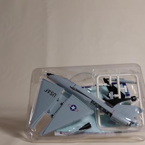 May include: A light gray model airplane with the text "USAF" on the wing, packaged in a clear plastic container. The model has a star and stripes emblem and is partially assembled.