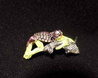 May include: A small, detailed figurine of a creature with a reptilian head and body, holding a fish. The creature has a brown, textured body, a red and white head, and yellow limbs. The fish is silver and the background is black.