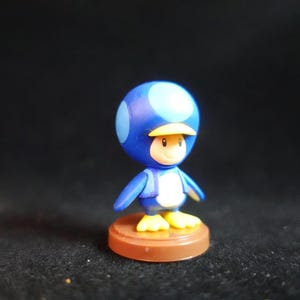 May include: A blue and white figurine of a character with a mushroom-shaped head, yellow feet, and a yellow visor. The figure is standing on a brown circular base. The character has a smiling face.