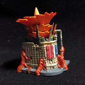 May include: A miniature, detailed sculpture of a building with a fiery orange top and a textured, beige facade. Red figures appear to be climbing the structure. The building has a section painted with red, white, and blue stripes, resembling a flag.