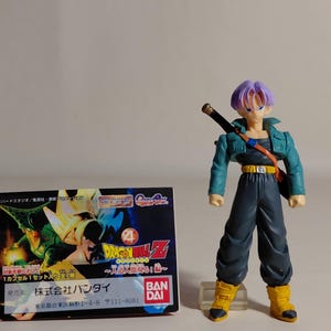 May include: A Dragon Ball Z action figure of Trunks, with purple hair, a teal jacket, and dark pants. A sword is strapped across his back. A small box with Japanese text and Dragon Ball Z graphics is to the left.
