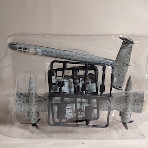 May include: A model airplane kit, featuring a gray and green camouflage design. The kit includes various unassembled parts, including wings, a fuselage, and a tail, all within a clear plastic package. The model appears to be a vintage aircraft.