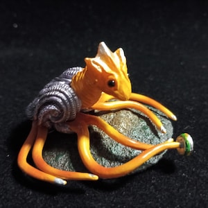 May include: A miniature sculpture of a creature with an orange body, tentacles, and a silver shell-like structure. The creature rests on a small, textured, gray rock. The sculpture has a detailed, handcrafted appearance.