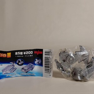 May include: A silver toy robot in a plastic bag next to a Japanese package with images of toy vehicles. The package has Japanese text and the words "FIRE STORM" in English. The toy robot is unassembled.