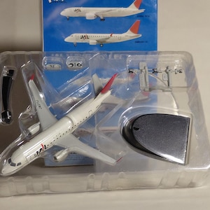 Può includere: Un modello di aereo JAL bianco con dettagli rossi sulla coda e sulle ali, esposto in una confezione di plastica trasparente. Il modello è dettagliato con il logo JAL e le marcature dell'aereo. Una scatola blu con immagini di aerei è visibile sullo sfondo.