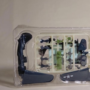 Puede incluir: Un kit de modelo de avión con varias piezas sin montar en un recipiente de plástico transparente. Las piezas son principalmente azul oscuro y verde, incluyendo alas, un fuselaje y otros componentes. Un ala tiene una estrella blanca.