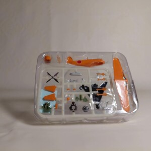 以下が含まれることがあります： オレンジ色の飛行機模型キット。翼、プロペラ、胴体など様々なパーツが透明なプラスチック容器に入っています。モデルには白い点のある赤い円があり、パーツは組み立て用に配置されています。