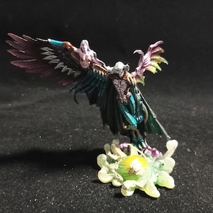 Puede incluir: Figura de fantasía en miniatura de una criatura alada con torso humanoide. La figura tiene una combinación de colores oscuros con acentos morados, verde azulado y amarillos. Está montada sobre una base floral, sobre un fondo oscuro.