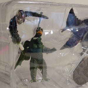 May include: A green and brown monster figurine with a bird-like head and wings, packaged in clear plastic. The figure is posed with outstretched arms and is accompanied by other dark blue and green pieces.