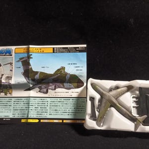Könnte beinhalten: Ein Modellflugzeug in einer weißen Kunststoffschale mit detailliertem Tarnmuster. Das Modell wird von einem gedruckten Informationsblatt mit japanischem Text und Flugzeugillustrationen begleitet. Das Modellflugzeug ist etwa 15 cm lang.