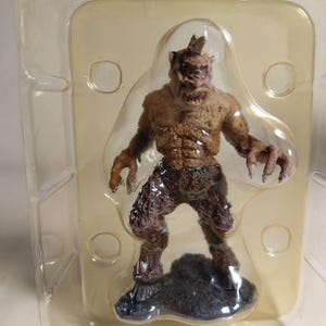 Peut inclure: Figurine détaillée d'un monstre avec un corps brun texturé, des griffes acérées et une tête cornue. La figurine est posée sur une base sombre et texturée et est enfermée dans un emballage en plastique transparent. Le monstre a une expression menaçante.