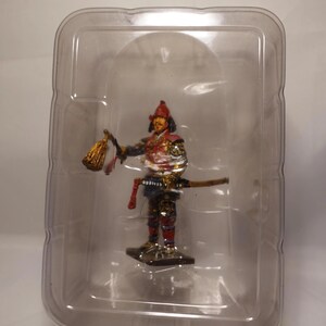May include: A miniature samurai figurine in a clear plastic case. The figure is dressed in detailed armour with red, gold, and blue accents. It holds a gold lantern and a sword. The figurine is standing on a small, black base.