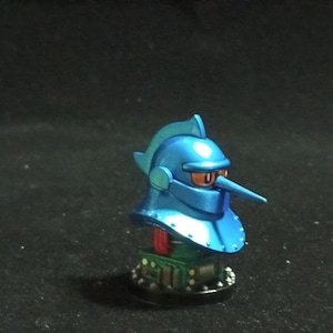 Puede incluir: Figura azul metálico de un casco de caballero con una nariz larga y puntiaguda y ojos naranjas. El casco se asienta sobre una base verde y roja con una parte inferior circular negra. Es un artículo decorativo único.