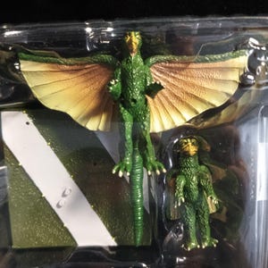 May include: A green and yellow winged creature figurine with a smaller matching figure. The larger figure has outstretched wings and is standing on a green and white angled platform. The figures are in a clear plastic package.