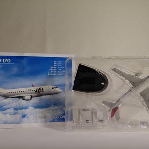 Può includere: Un modello di aereo Embraer 170 bianco e rosso in una custodia di plastica trasparente. Il modello presenta il logo JAL ed è esposto con un supporto. La confezione include il testo "EMBRAER 170".