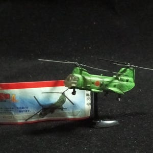 May include: A green toy helicopter with a red and white emblem, displayed on a black stand. A colorful product label is visible to the left. The helicopter has two rotors and a detailed design.