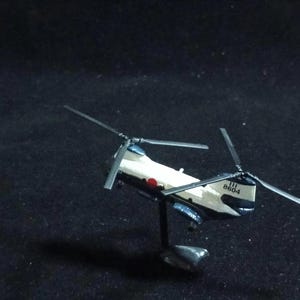 May include: A miniature model helicopter with a white and navy blue body, featuring a red circle and black text. The helicopter has two sets of blades and is mounted on a black stand. The model is set against a dark background.
