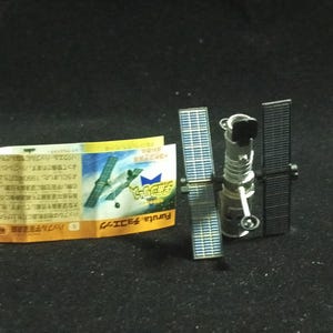 May include: A miniature model of the Hubble Space Telescope with solar panels and a cylindrical body. Beside it is a folded paper with Japanese text and an illustration of a satellite. The model is silver and black.
