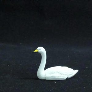 May include: A small, white swan figurine with a yellow beak. The swan is depicted in a resting position, with its neck gracefully curved. The figurine is set against a dark background, highlighting its form.