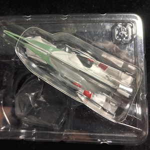 May include: A small, white and silver toy airplane with red and green accents is encased in a clear plastic package. The airplane has a sleek design, with wings and a pointed nose. The packaging is rectangular and transparent.