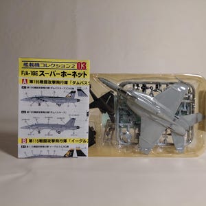 以下が含まれることがあります： F/A-18Eスーパーホーネットの模型キット。キットは透明なプラスチック容器に入っており、未組み立ての灰色のプラスチック部品が見えます。日本語のテキストと飛行機のイラストが描かれた箱がキットの隣にあります。