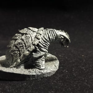 May include: A detailed, silver-colored figurine of a fantasy creature, possibly a dragon or dinosaur. The figure has textured scales and a detailed head, mounted on a circular base. The creature is in a crouching position.