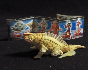 Può includere: Una statuetta di dinosauro color beige e crema con un corpo strutturato e strisce viola sulla schiena. Un piccolo pezzo giallo traslucido è vicino al dinosauro. Sullo sfondo, un foglio di carta piegato con immagini di altre figure.
