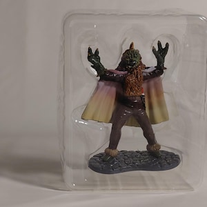 May include: A figurine of a creature with a green head, brown beard, and a cape with yellow and pink hues. The figure is in a clear plastic container, standing on a dark blue base.